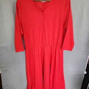 May include: A bright red knee-length dress featuring three-quarter length sleeves, a round neckline, and a partial button-down placket at the center chest. The garment has a gathered elasticized waistline and a simple, solid-colored fabric design.
