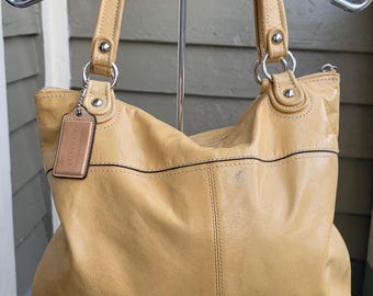 Peut inclure: Un sac cabas en cuir jaune moutarde doté de deux bandoulières, de finitions en métal argenté, d'une fermeture éclair sur le dessus et d'une étiquette en cuir rectangulaire gravée du mot COACH sur une chaîne argentée.
