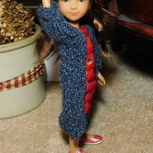 Puede incluir: Una muñeca con un gorro rojo tejido con detalles coloridos, un abrigo azul tejido y un vestido rojo tejido. La muñeca también lleva zapatos de purpurina rosa. La muñeca está de pie sobre una alfombra beige.