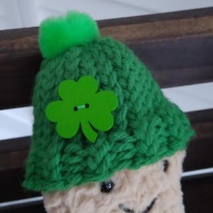 Può includere: Un arachide di peluche che indossa un cappello di lana verde con un bottone a forma di trifoglio. Il cappello ha un pon pon verde in cima.