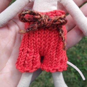 Puede incluir: Muñeca hecha a mano con cuerpo y extremidades de tela beige. La muñeca lleva un pantalón corto de punto rojo brillante con un cinturón marrón y beige. Los brazos de la muñeca están extendidos y tiene una pequeña cola blanca.