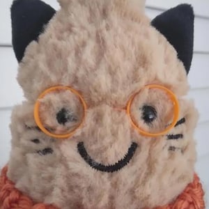 Könnte beinhalten: Ein Plüschtier in Katzenform mit beigem, flauschigem Fell, schwarzen Ohren und einer runden, orangefarbenen Kunststoffbrille. Die Katze hat ein aufgesticktes schwarzes Lächeln und Schnurrhaare. Sie trägt einen dicken, orange-weißen Häkelkragen.