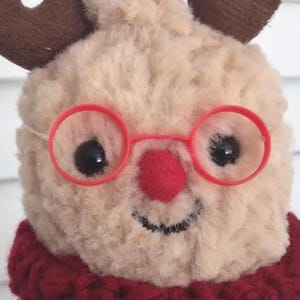 May include: A plush reindeer figure made of beige yarn with brown felt antlers, a red pom-pom nose, and small black eyes. The figure wears round red plastic glasses and a knitted sweater featuring a red and white chevron pattern with a solid red collar.