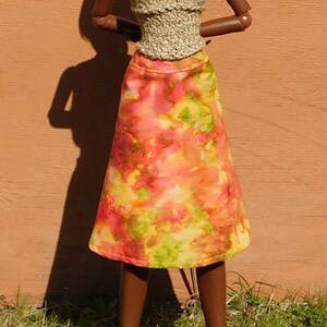 May include: A doll wearing a gold crochet top and a tie-dye skirt with orange, yellow, and green colors. The doll has brown skin and black hair. The doll is wearing brown shoes.