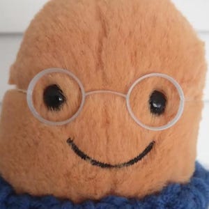 May include: A plush toy with a tan, fuzzy, rounded head featuring black embroidered eyes and a simple curved smile. The figure wears thin, clear plastic circular glasses and a blue and white crocheted sweater with a textured, ribbed collar.