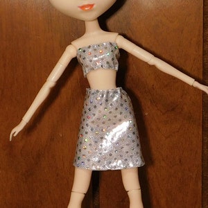 May include: A doll with blue eyes and a blue knitted beret is wearing a silver sequined skirt and a matching top.
