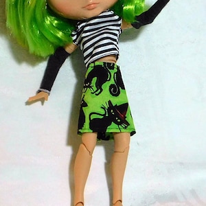 May include: A green haired doll with green eyes wearing a black and white striped shirt and a green skirt with black cats printed on it. The doll is wearing white shoes and has a friendly expression.