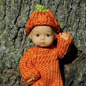 May include: A knitted orange pumpkin costume for a doll. The costume includes a hat and a jumpsuit. The doll is wearing the costume and is standing in front of a tree.