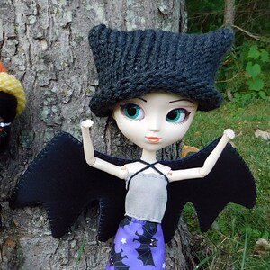Può includere: Una bambola con ali nere e una gonna viola con pipistrelli. La bambola indossa un cappello nero di lana con orecchie da gatto e ha grandi occhi blu. La bambola è in piedi davanti a un albero.