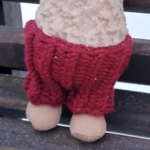 Peut inclure: Une peluche beige avec un visage souriant et des yeux noirs. Elle porte un pantalon rouge tricoté et a des pieds beiges. Le jouet est assis sur un banc en bois.