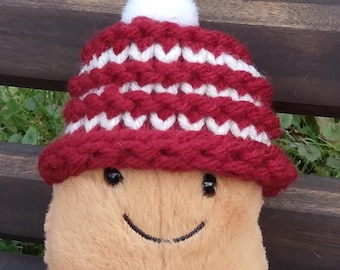 Puede incluir: Un juguete de peluche con forma de patata que lleva un gorro de punto rojo y blanco con un pompón blanco en la parte superior. La patata tiene una cara sonriente y piernas de pana marrón.
