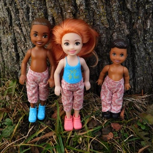 May include: Three dolls are posed in front of a tree trunk. The dolls have brown skin and wear patterned pants. One doll wears a blue tank top with the text "Just Be You". The dolls wear blue, pink, and black shoes.