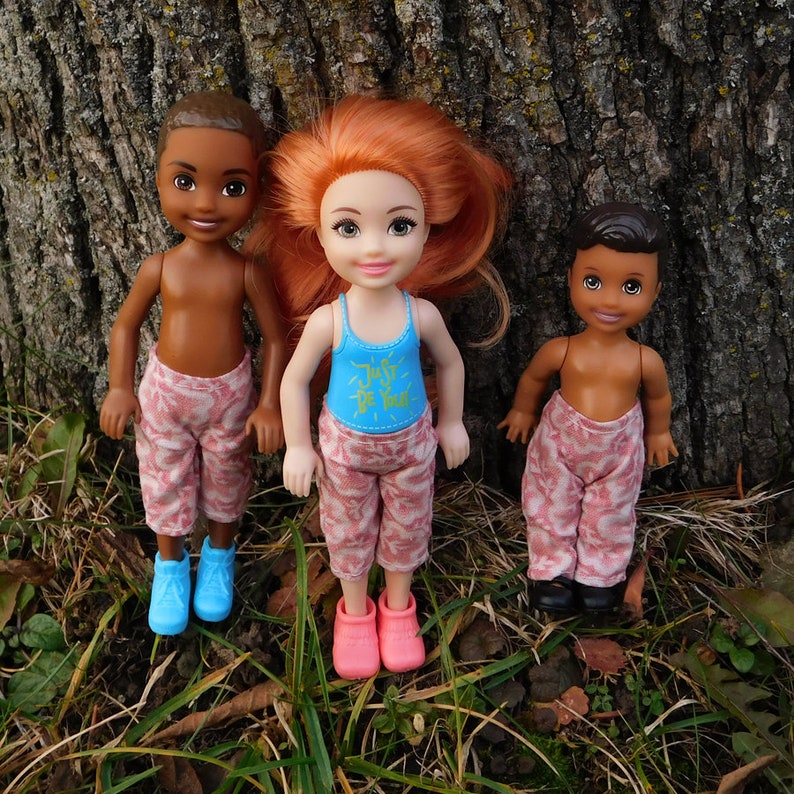 May include: Three dolls are posed in front of a tree trunk. The dolls have brown skin and wear patterned pants. One doll wears a blue tank top with the text "Just Be You". The dolls wear blue, pink, and black shoes.