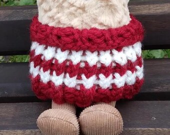 Puede incluir: Un juguete de peluche con una falda de crochet roja y blanca. El juguete está sentado en un banco de madera.