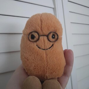 Puede incluir: Un juguete de peluche con forma de jud&iacute;a, en tonos naranja y marr&oacute;n. El juguete tiene unas gafas peque&ntilde;as y redondas con montura de alambre y una sonrisa cosida. Los pies son de pana. El juguete est&aacute; dise&ntilde;ado para ser sujetado.