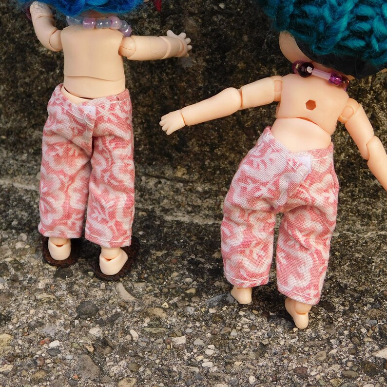 May include: Two doll figures wearing pink and white patterned pants and brown sandals. The dolls have blue and teal yarn hair and beaded necklaces. The dolls are posed with their backs to the viewer.
