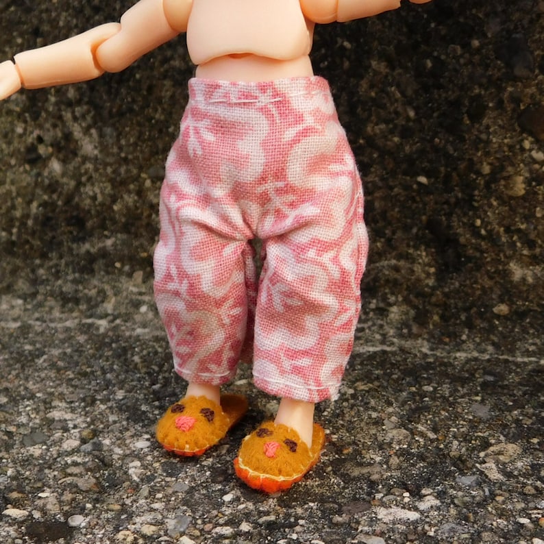 May include: Doll-sized pink and white patterned pants with matching slippers. The pants are a cropped style, and the slippers are orange and brown with a face design. The doll's skin is a light tan color.