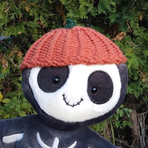 May include: A plush toy with a skeleton design wearing a knitted pumpkin hat. The toy has a white face with black eyes and a stitched smile.