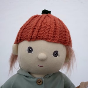 May include: A handmade doll with a knitted pumpkin hat, wearing a light green hooded jacket. The doll has a light beige face with embroidered eyes and a button nose.