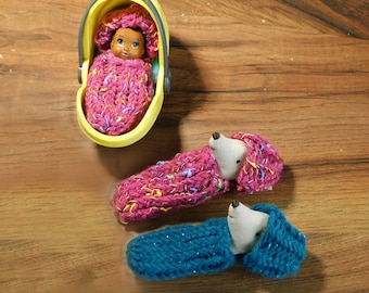 May include: Three knitted sleeping bags for dolls. One is pink with white and yellow flecks, one is teal, and one is pink with white and yellow flecks. The pink sleeping bag is in a yellow plastic doll carrier.