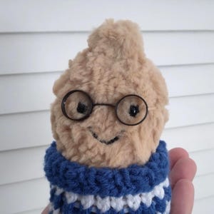 Puede incluir: Una figura de peluche marr&oacute;n claro con gafas redondas con montura de alambre y una sonrisa cosida. Lleva un cuello de ganchillo a rayas azules y blancas. La figura se sostiene en una mano sobre un fondo blanco.