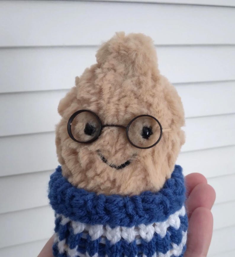 Puede incluir: Una figura de peluche marr&oacute;n claro con gafas redondas con montura de alambre y una sonrisa cosida. Lleva un cuello de ganchillo a rayas azules y blancas. La figura se sostiene en una mano sobre un fondo blanco.