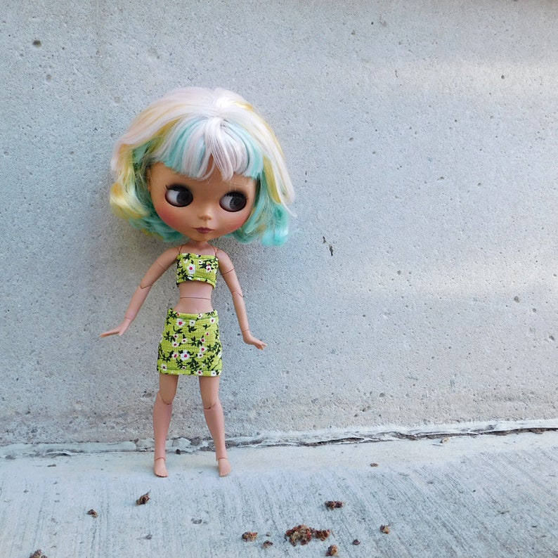 May include: A doll with light blue and blonde hair wearing a green and white floral print crop top and skirt.
