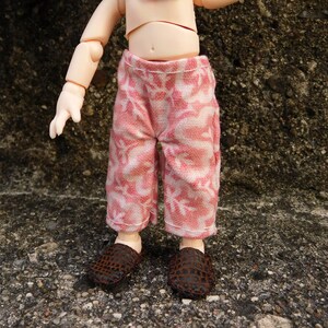May include: Doll-sized pink and white patterned pants with matching brown woven shoes. The pants have a floral design and are cropped at the ankles. The shoes have a woven pattern.