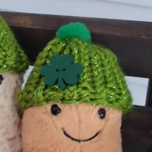 Può includere: Due pupazzi con facce sorridenti, che indossano cappelli di maglia verdi con un trifoglio e un pompon verde. I cappelli hanno un filo metallico intrecciato nel filato verde. Le figure hanno un colore beige chiaro e arancione.