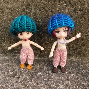 May include: Two small dolls are shown, each wearing a knitted hat. One hat is teal, the other is blue. Both dolls wear pink patterned pants and small shoes. One doll has orange shoes, the other has brown shoes. Both dolls have necklaces.