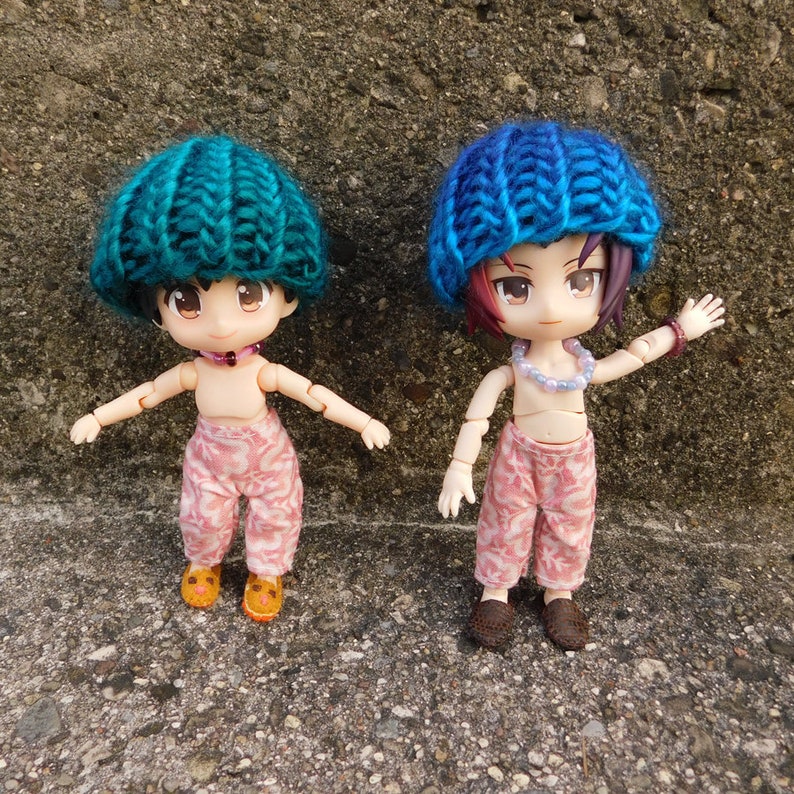 May include: Two small dolls are shown, each wearing a knitted hat. One hat is teal, the other is blue. Both dolls wear pink patterned pants and small shoes. One doll has orange shoes, the other has brown shoes. Both dolls have necklaces.