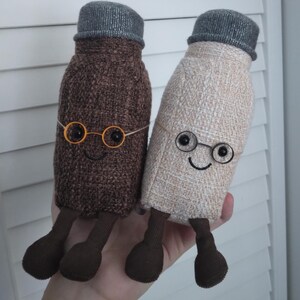 Puede incluir: Dos mu&ntilde;ecos de peluche con forma de botella con caras sonrientes, gafas redondas peque&ntilde;as y sombreros grises. Uno es marr&oacute;n, el otro beige. Ambos tienen piernas y pies marrones. Los mu&ntilde;ecos son hechos a mano y caprichosos.