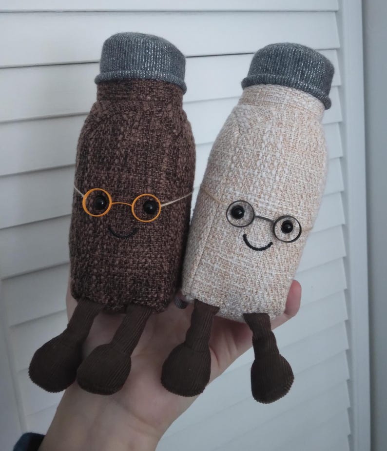 Puede incluir: Dos mu&ntilde;ecos de peluche con forma de botella con caras sonrientes, gafas redondas peque&ntilde;as y sombreros grises. Uno es marr&oacute;n, el otro beige. Ambos tienen piernas y pies marrones. Los mu&ntilde;ecos son hechos a mano y caprichosos.
