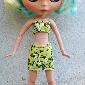 May include: A green and black floral print crop top and skirt set for a doll. The top is strapless and the skirt is a mini skirt.