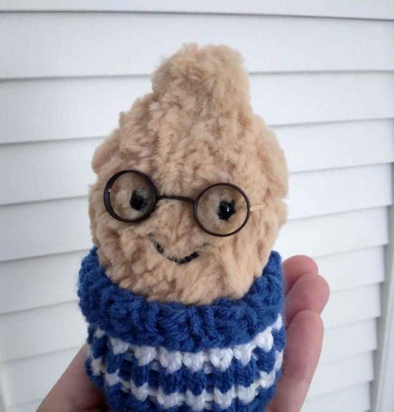 Puede incluir: Un juguete de peluche en forma de una figura difusa de color marr&oacute;n claro con gafas redondas negras y una cara sonriente. Lleva un su&eacute;ter a rayas azules y blancas. El juguete se sostiene en una mano.