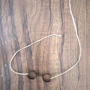 Puede incluir: Gafas peque&ntilde;as y redondas con montura negra y una cuerda blanca adjunta. Las gafas est&aacute;n sobre una superficie de madera. Las gafas tienen un dise&ntilde;o simple y minimalista.