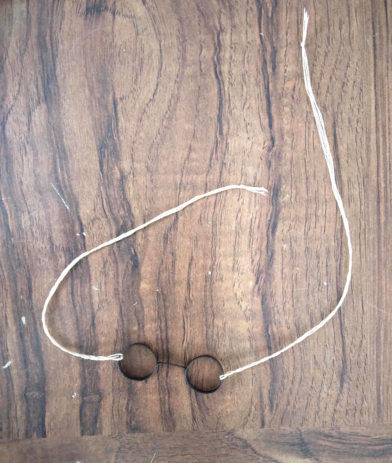 Puede incluir: Gafas peque&ntilde;as y redondas con montura negra y una cuerda blanca adjunta. Las gafas est&aacute;n sobre una superficie de madera. Las gafas tienen un dise&ntilde;o simple y minimalista.