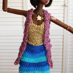 Puede incluir: Una muñeca con un atuendo de ganchillo hecho a mano. El atuendo incluye un top dorado, una falda a rayas azules y turquesas y un chaleco morado. Un colgante de tortuga blanca cuelga de un collar gris.