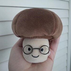 Puede incluir: Un juguete de peluche con forma de hongo con un sombrero marr&oacute;n y un tallo color crema. El juguete tiene gafas con montura negra y una cara sonriente. El hongo se sostiene en una mano.