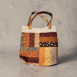 May include: A tote bag featuring a patchwork design with various fabric patterns in shades of orange, brown, burgundy, and cream. The bag has two matching fabric handles and a rectangular body constructed from vertical and horizontal fabric strips.