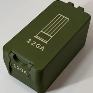 May include: An olive green rectangular storage box with a hinged lid. The top features a white line-art icon of a shotgun shell and the text 12GA. The front latch also displays the text 12GA in white. The box has a matte finish.