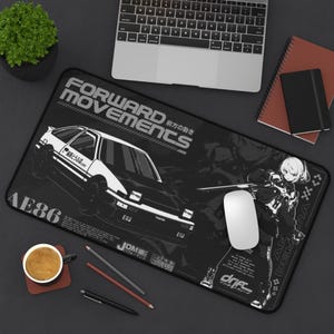 以下が含まれることがあります： AE86の車の白いグラフィック、FORWARD MOVEMENTSの文字、アニメスタイルのキャラクターが描かれた黒い長方形のデスクマット。マットには日本語の文字、JDMという単語、ドリフト関連のブランドロゴが暗い背景にデザインされています。