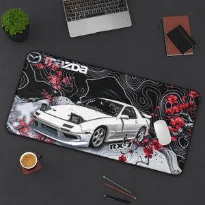 Pode incluir: Um tapete de secretária retangular com um gráfico de um carro desportivo Mazda RX-7 branco sobre um fundo preto com linhas topográficas brancas e ramos de cerejeira vermelhos. O texto Mazda e RX-7 está impresso a branco.