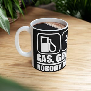 Könnte beinhalten: Ein weißer Keramikbecher mit einem schwarzen Grafikdesign, das ein Zapfsäulensymbol und ein Cannabisblattsymbol zeigt. Der auf den Becher gedruckte Text lautet GAS, GRASS, NOBODY RIDES FOR FREE in fetten weißen Großbuchstaben auf schwarzem Grund.