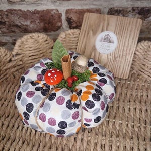 May include: A fabric pumpkin decoration featuring a white base with black, orange, and purple polka dots. The top is adorned with a cinnamon stick, a green leaf, a small red mushroom, an acorn, and green moss. A wooden plaque with the text Gem Cottage is behind it.