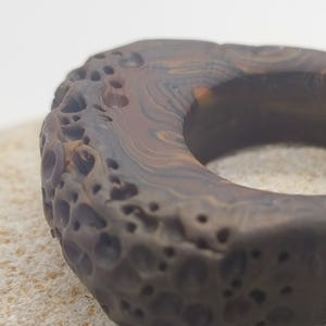 May include: A circular ring with a dark brown and black marbled pattern. The outer surface features a porous, cratered texture, while the inner surface is smooth. The object has a thick, rounded profile with visible swirling colour variations.