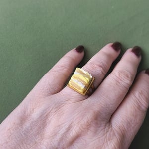 May include: A gold-toned ring featuring a square-shaped charm designed to resemble a layered slice of cake. The charm displays horizontal stripes in shades of yellow, white, and light brown, mounted on a simple metallic band.