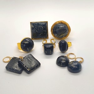 Puede incluir: Una colección de joyería con piezas de resina veteada en azul oscuro y dorado montadas en metal de tono dorado. El conjunto incluye tres anillos de diversas formas, un par de pendientes cuadrados y un par de pendientes redondos con aros.