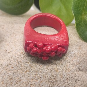 May include: A bright red ring featuring a smooth, thick band and a cluster of small, rounded, bubble-like protrusions on the front surface. The entire piece has a glossy finish and a uniform, vibrant red colour throughout its solid construction.