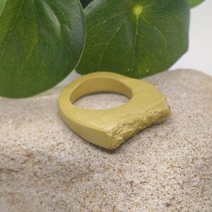 Puede incluir: Un anillo de color amarillo mostaza con una banda lisa y redondeada y una sección texturizada e irregular en un lado. El anillo presenta un acabado mate y una construcción sólida y gruesa, mostrando un contraste entre las superficies.